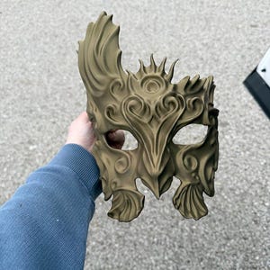 Può includere: Una maschera verde oliva dettagliata con un design ornato. La maschera presenta motivi a spirale, bordi appuntiti e ritagli per gli occhi. La maschera è tenuta su uno sfondo grigio.