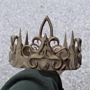 Può includere: Una corona color oro con dettagli ornati. La corona presenta una serie di punte rivolte verso l'alto e decorazioni. Il design include una forma centrale a giglio stilizzata. La corona sembra essere fatta di un materiale solido.