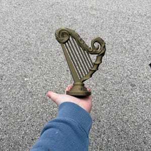 Può includere: Una statuetta a forma di arpa color bronzo con volute decorative nella parte superiore e inferiore. Le corde dell'arpa sono visibili e la statuetta è tenuta in mano su uno sfondo grigio. L'arpa è un oggetto decorativo.