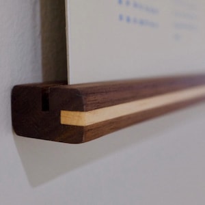 May include: A wooden poster hanger with a dark brown finish and a light wood accent. The hanger is designed to hold a poster or print, and is mounted on a white wall. The design is minimalist and modern.