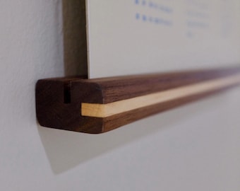 May include: A wooden poster hanger with a dark brown finish and a light wood accent. The hanger is designed to hold a poster or print, and is mounted on a white wall. The design is minimalist and modern.