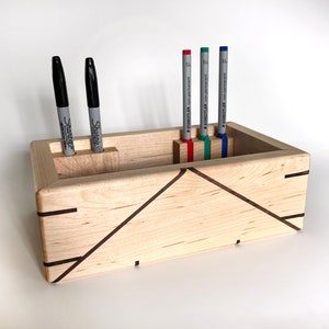 May include: A wooden pen holder with a geometric design. The holder has four slots for pens and is made of light wood with dark wood accents. There are four pens in the holder, two black Sharpie pens and two blue and green highlighters.