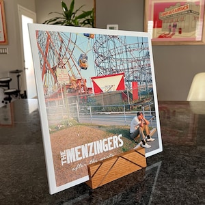 May include: A white vinyl record with a photo of a roller coaster and a person sitting on a bench. The record is titled "The Menzingers - After the Party". The record is displayed on a wooden record stand.