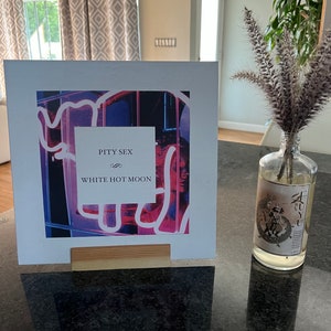 Könnte beinhalten: Weißes Albumcover mit einem neonrosa und blauen Design. Der Albumtitel lautet "Pity Sex" und der Bandname ist "White Hot Moon".