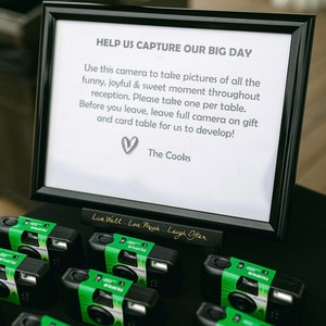 May include: A black framed sign with the text "HELP US CAPTURE OUR BIG DAY" and a heart symbol. Below are several black Fujifilm cameras with green labels. The sign also says "Live Well - Love Much - Laugh Often".