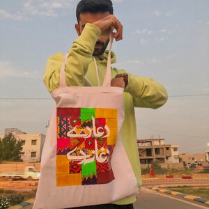 May include: A white tote bag with a colorful patchwork design featuring Arabic calligraphy. The bag has a green, red, yellow, and blue color scheme.