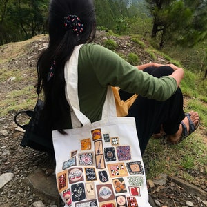 May include: A white tote bag with a colorful design featuring various musical instruments and patterns. The bag is decorated with Urdu text and is sitting on a rocky surface.