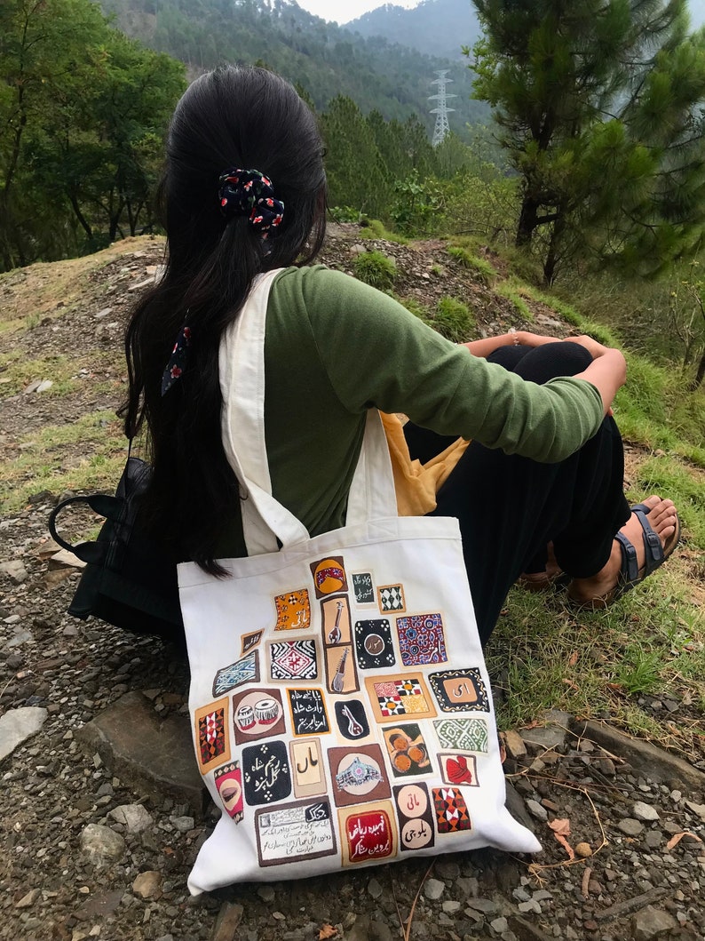May include: A white tote bag with a colorful design featuring various musical instruments and patterns. The bag is decorated with Urdu text and is sitting on a rocky surface.