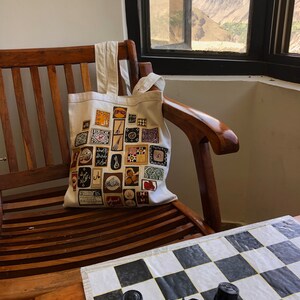 May include: A white canvas tote bag with a colorful design featuring various patterns and Arabic script. The bag is sitting on a wooden chair with a chessboard on a table in the foreground.