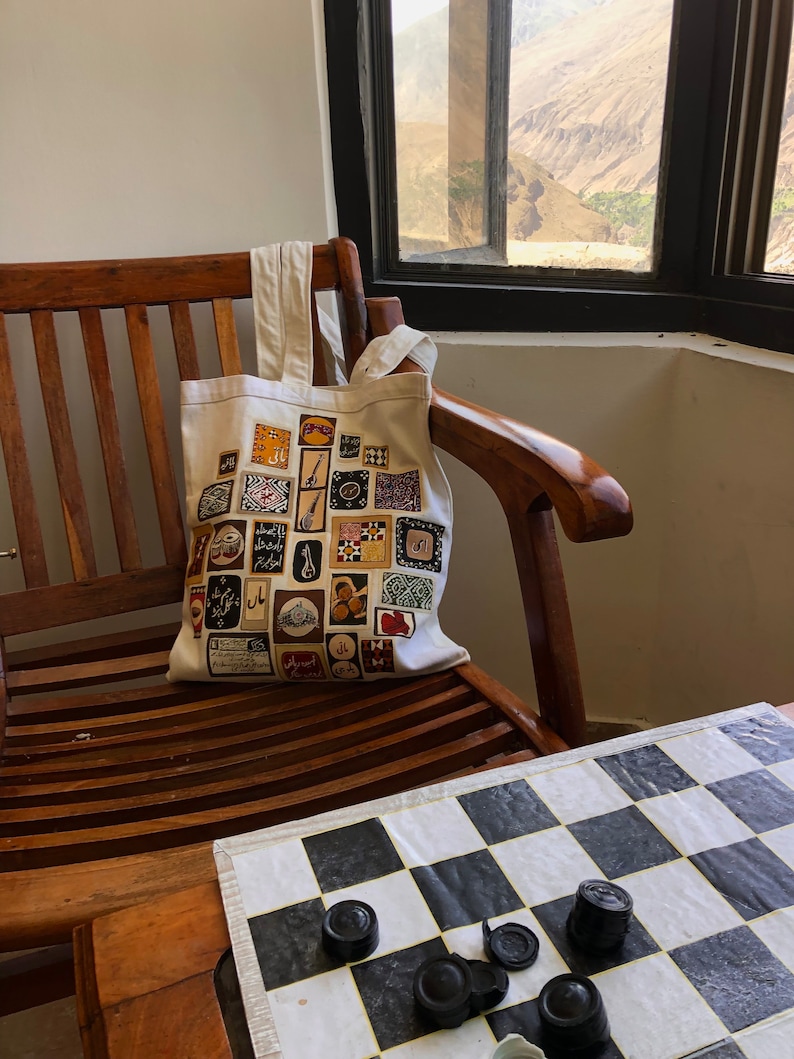 May include: A white canvas tote bag with a colorful design featuring various patterns and Arabic script. The bag is sitting on a wooden chair with a chessboard on a table in the foreground.