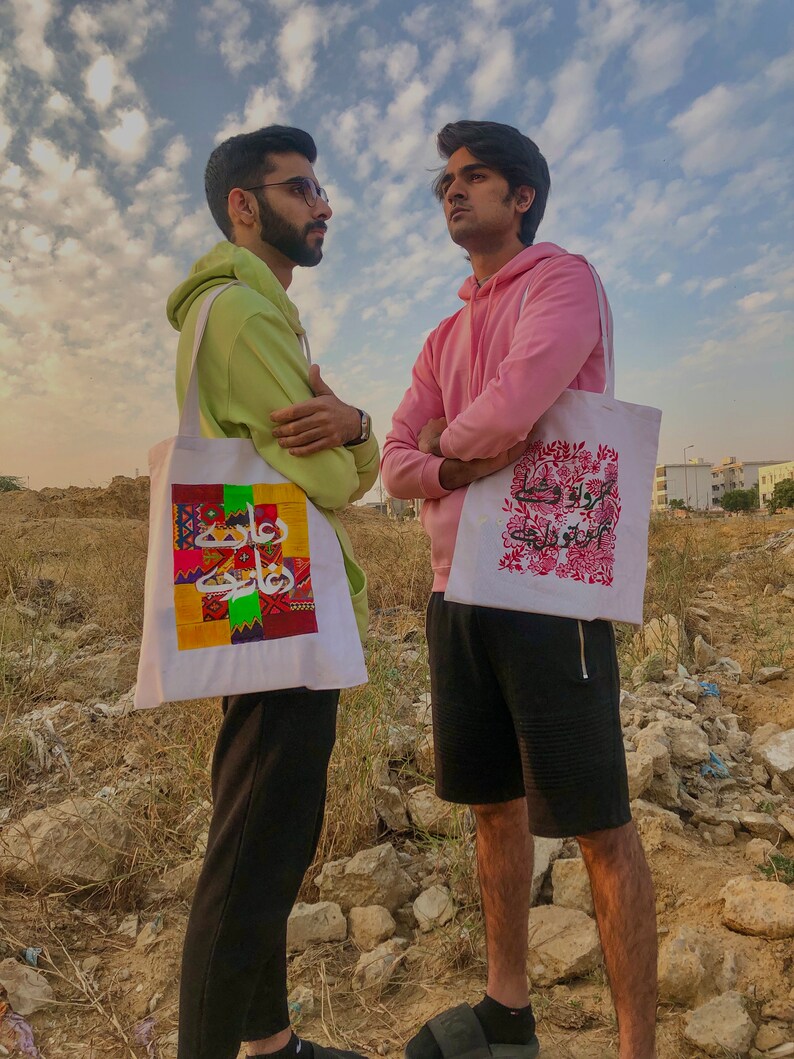 May include: Two people wearing hoodies are standing outdoors, each carrying a white tote bag with colorful designs. The person on the left is wearing a light green hoodie and the person on the right is wearing a pink hoodie. The tote bag on the left has a colorful geometric design with Arabic script. The tote bag on the right has a floral design with Arabic script.