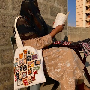 May include: A woman wearing a black headscarf sits on a chair and reads a book. She is carrying a white tote bag with a colorful patchwork design. The bag is decorated with various patterns and symbols, including geometric shapes, flowers, and Arabic script.