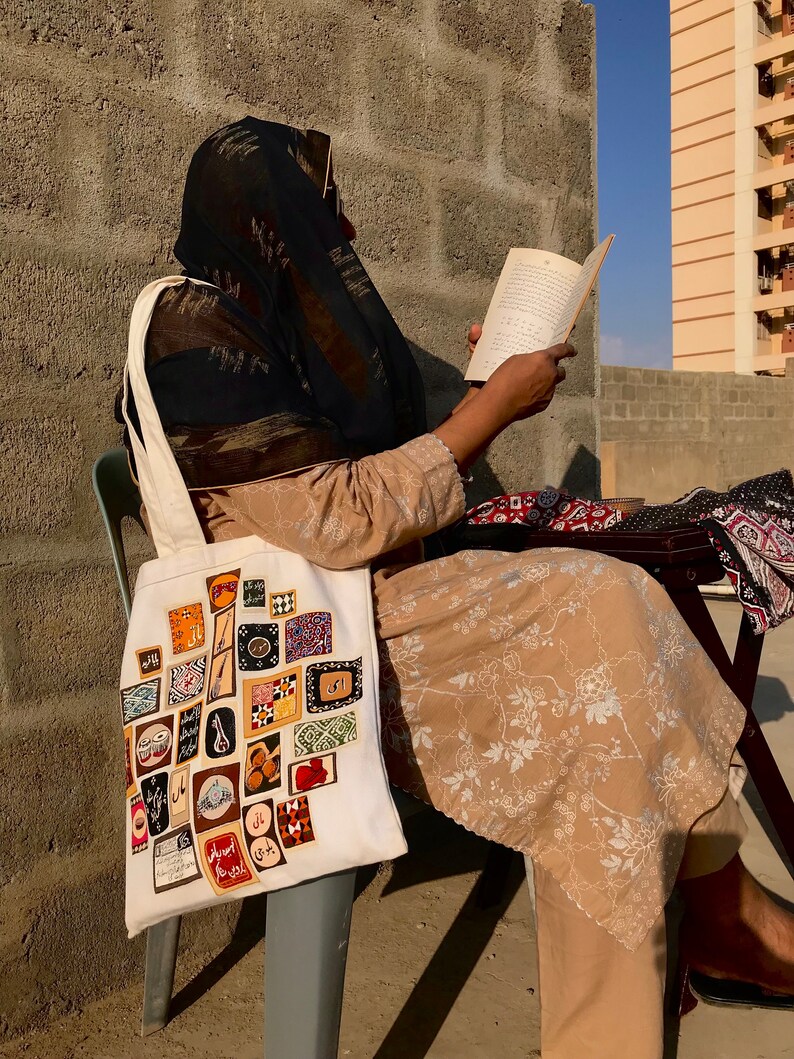 May include: A woman wearing a black headscarf sits on a chair and reads a book. She is carrying a white tote bag with a colorful patchwork design. The bag is decorated with various patterns and symbols, including geometric shapes, flowers, and Arabic script.