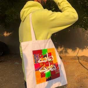 May include: A white tote bag with a colorful design featuring Arabic calligraphy and geometric patterns. The bag has a green, red, yellow, and blue color scheme.