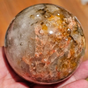 May include: A clear quartz sphere with inclusions of brown, orange, and white. The sphere is polished and has a smooth surface.