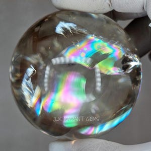 Può includere: Una sfera di cristallo trasparente con iridescenze arcobaleno interne. La sfera è tenuta da una mano guantata. Il testo "JLR RADIANT GEMS" è visibile sul fondo della sfera.