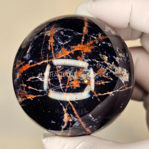 Könnte beinhalten: Eine polierte Kugel mit dunkelblauer Grundfarbe, versehen mit orangefarbenen und weißen, adernartigen Mustern. Die Kugel hat eine glänzende Oberfläche und der Text "RADIANT GEMS LLC" ist sichtbar.