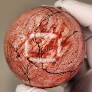 Puede incluir: Una esfera de piedra de color marrón rojizo con vetas oscuras y una textura agrietada. La piedra está sujeta por una mano enguantada. La esfera tiene una superficie pulida y un aspecto natural y terroso.