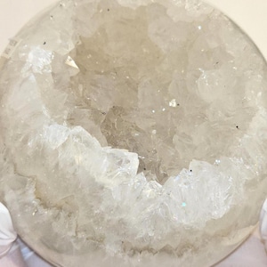 May include: A round, geode-like object with a crystalline interior. The interior features a cluster of clear, pointed crystals. The outer shell is a translucent, off-white color. The crystals sparkle with hints of iridescence.