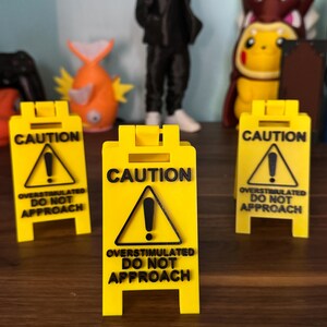 May include: Three yellow caution signs with black text and a black exclamation point inside a black triangle. The text on the signs reads "Caution Overstimulated Do Not Approach".