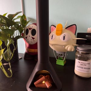 May include: A black ceramic fireplace with a glowing orange light inside. The fireplace is on a dark wood surface with a small jar of candle and a figurine of a cartoon character.