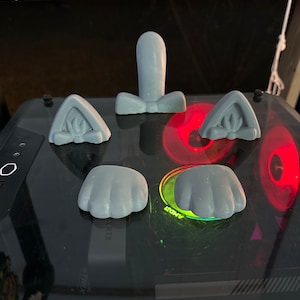 May include: Five grey 3D printed cat ears and paws. The ears are pointed and the paws have four toes. The objects are on a black surface with a red and green light.