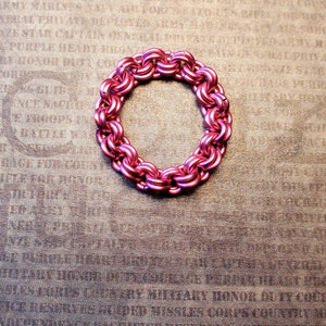 May include: A pink metal chain bracelet with a circular design. The bracelet is lying on a brown textured surface.