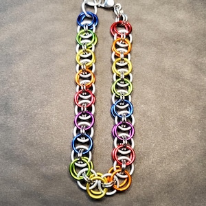 May include: A chainmail bracelet with a rainbow colour scheme. The bracelet is made of small, brightly coloured metal rings, including red, orange, yellow, green, blue, and purple. It has a silver clasp and is shown against a dark grey backdrop.