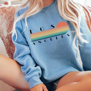 May include: A light blue sweatshirt with a retro-style graphic of a sunset and the text "LOS ANGELES" in black.