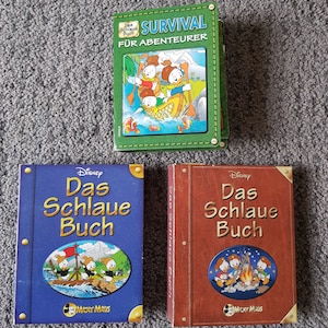 Könnte beinhalten: Drei Hardcover-Bücher mit blauen, roten und grünen Einbänden. Die Bücher zeigen Illustrationen von Donald Duck und seinen Neffen. Die Titel der Bücher lauten "Disney Das Schlaue Buch", "Disney Das Schlaue Buch" und "Survival Für Abenteurer".