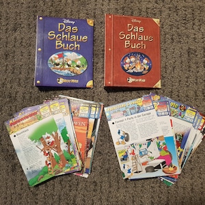 Könnte beinhalten: Zwei rote und blaue Hardcover-Bücher mit dem Titel "Disney Das Schlaue Buch" mit Mickey-Mouse-Illustrationen. Ein Stapel bunter Karten mit Illustrationen und Text auf Deutsch.