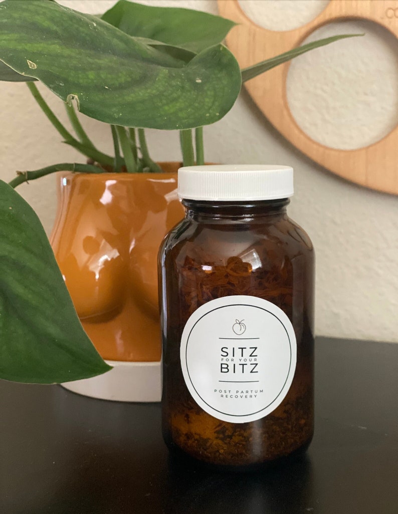 以下が含まれることがあります： 白い蓋と白いラベルが付いた茶色のガラス瓶。ラベルには「Sitz for your Bitz Post Partum Recovery」と書いてあります。瓶は茶色の表面の上に置かれています。