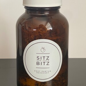 以下が含まれることがあります： 白い蓋が付いた茶色のガラス瓶に乾燥ハーブが入っています。瓶には、桃のアイコンと「Sitz for your Bitz Post Partum Recovery」というテキストが書かれた白いラベルが付いています。