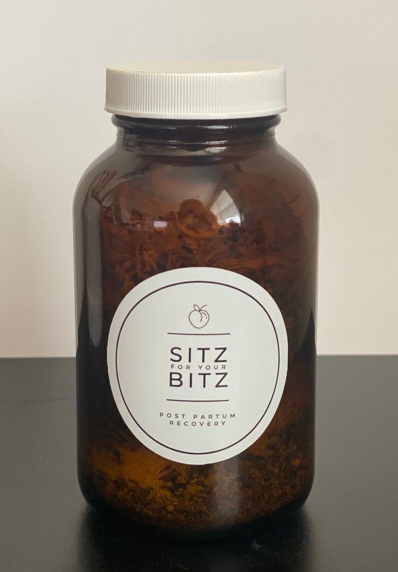 以下が含まれることがあります： 白い蓋が付いた茶色のガラス瓶に乾燥ハーブが入っています。瓶には、桃のアイコンと「Sitz for your Bitz Post Partum Recovery」というテキストが書かれた白いラベルが付いています。
