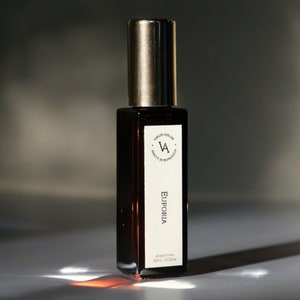 Peut inclure: Flacon de parfum rectangulaire contenant un liquide ambré foncé et un bouchon doré. L'étiquette indique "Eufória" et "oil perfume 10ml / 0.33oz". Le flacon présente un design minimaliste, avec le logo et le texte de la marque sur une étiquette blanche.