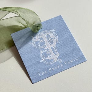 Könnte beinhalten: Ein hellblaues quadratisches Geschenk-Etikett mit einem weißen, verzierten Buchstaben "P" und dem Text "The Peake Family". Ein durchsichtiges grünes Band ist am Etikett befestigt.