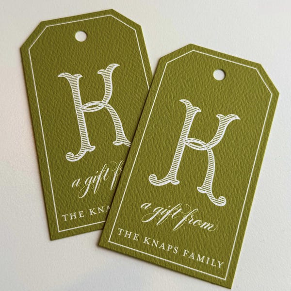 Puede incluir: Dos etiquetas de regalo verde oliva con borde blanco y letra "K" blanca. Las etiquetas dicen "a gift from THE KNAPS FAMILY". Cada etiqueta tiene un agujero en la parte superior para ensartar.