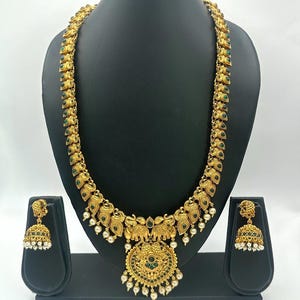May include: A gold-toned necklace featuring repeating leaf-shaped links with green accents, a central pendant with two elephant motifs, and small white pearl-like beads. Matching bell-shaped earrings with green and white bead details.