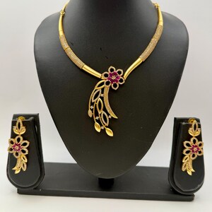 May include: A gold-toned necklace and earring set displayed on a black stand. The necklace features a floral pendant with a ruby-colored center and a flowing leaf design. The earrings mirror the floral design. The necklace has a textured band.