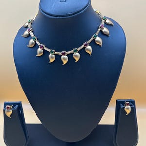 May include: A gold-toned necklace and matching stud earrings set. The necklace features a chain with alternating square-cut green and red stones, interspersed with dangling gold-toned leaf-shaped charms. The earrings feature a single red stone.