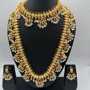 Puede incluir: Un conjunto de joyas de color dorado que incluye un collar, un collar corto tipo gargantilla y pendientes a juego. Las piezas están adornadas con detalles rojos, verdes y blancos, y pequeñas perlas blancas. El conjunto se exhibe en un expositor de joyas negro.