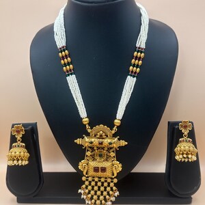 May include: A gold-toned necklace and earring set displayed on a black mannequin. The necklace features a pendant with red and green accents, and a white beaded strand. The earrings are bell-shaped with floral details. The jewelry set is ornate and detailed.