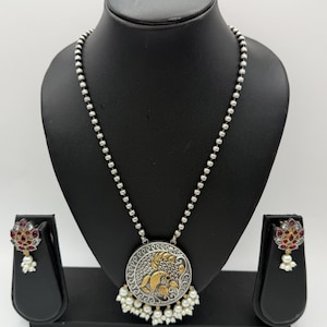 May include: A silver-toned necklace and earring set. The necklace features a round pendant with a gold and silver design, and a beaded chain. The earrings have a floral design with red and white accents, and a pearl drop.