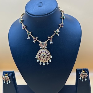 May include: A gold-toned necklace and earring set featuring a circular pendant with red and green stones, accented by small white pearls. The necklace chain includes alternating square red and green stone links, while the matching earrings feature a floral-inspired design with similar stones and pearl drops.