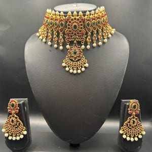 May include: A gold-toned choker necklace and matching earrings set. The necklace features red and green gemstones, with dangling pearl accents. The earrings mirror the necklace design. The jewelry is displayed on a black mannequin.