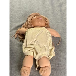 May include: A doll with a soft, tan-coloured fabric body and head, featuring light brown yarn hair. The doll is wearing a sleeveless, pale yellow romper with a single visible button at the bottom hem. The facial features include a small pink mouth.