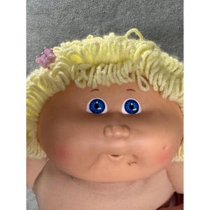 May include: A doll with a round, peach-toned vinyl face, bright blue eyes, and a small nose. The doll has pale yellow yarn hair styled in curls, accented by a small, translucent pink butterfly-shaped hair clip on the left side.
