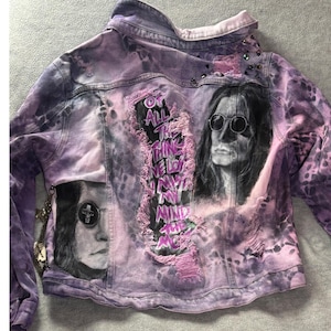 Puede incluir: Una chaqueta vaquera con estampado tie-dye en morado y negro que presenta dos retratos en blanco y negro de una persona con gafas redondas. Un panel central desgastado muestra el texto "OF ALL THE THINGS I LOVE I MISS MY MIND THE MOST" en morado.