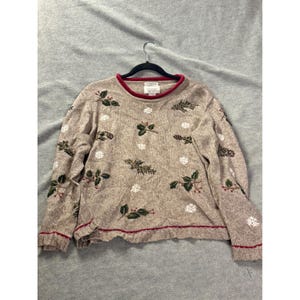 May include: A beige long-sleeve sweater featuring a pattern of embroidered green holly leaves with red berries and white snowflakes. The sweater has a crew neckline with a red trim, a matching red trim at the hem, and a white label at the inner collar.