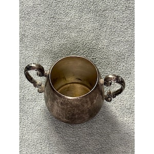May include: A small, tarnished silver-toned metal sugar bowl with a rounded body and two ornate, curved handles on opposite sides. The interior of the bowl shows a smooth, metallic finish, while the exterior surface displays a weathered patina.